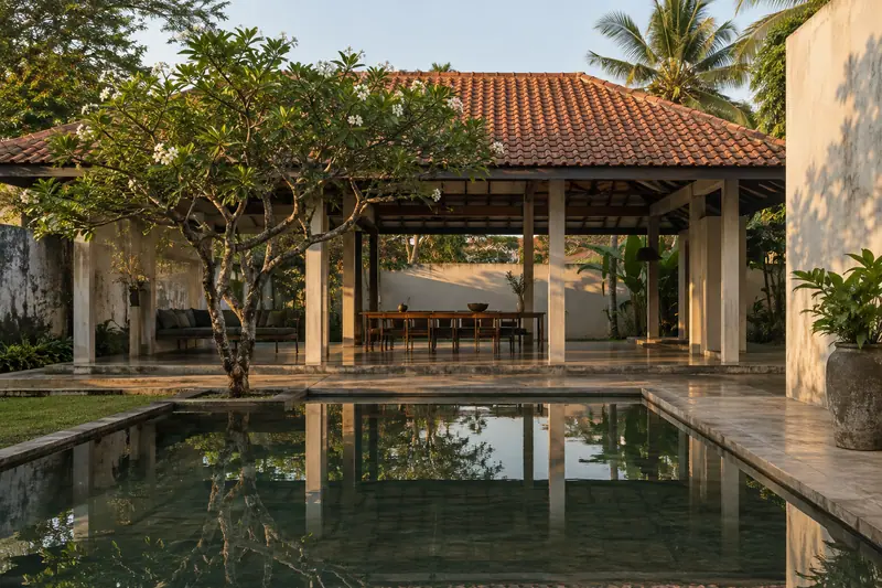 A clay-tiled courtyard pavilion with slender columns, a frangipani tree and a reflecting pool in late afternoon light.