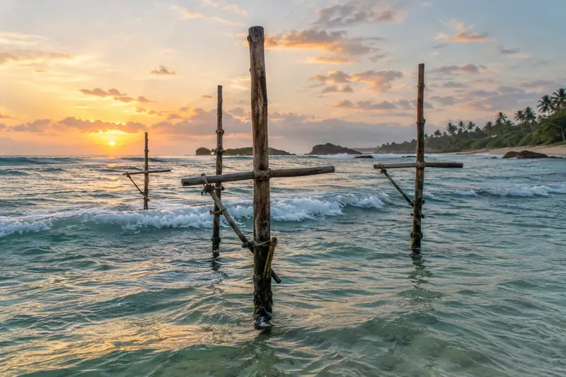Empty wooden fishing stilts with crossbars stand in turquoise surf at sunrise beside a palm-lined beach.
