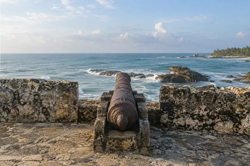A rusted iron cannon on a weathered coral-stone rampart overlooks the Indian Ocean in soft morning light.