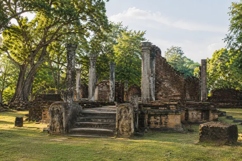 Weathered brick ruins and carved granite pillars stand in short grass beneath large trees in late afternoon light.