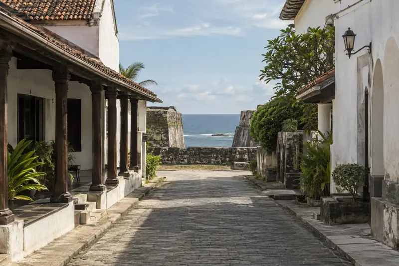 A quiet cobbled lane between whitewashed buildings with terracotta roofs leads to stone ramparts overlooking the sea.