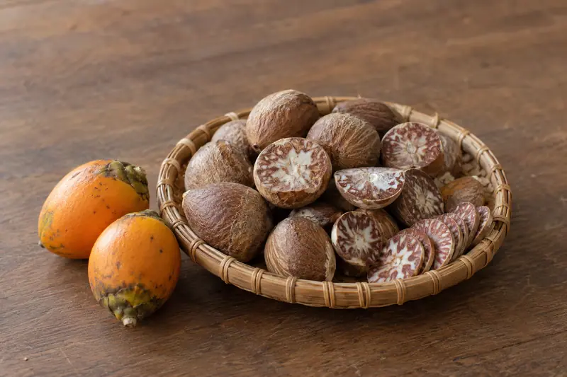 Whole and sliced dried areca nuts in a woven cane basket beside two orange-husked fruits on a wooden table.