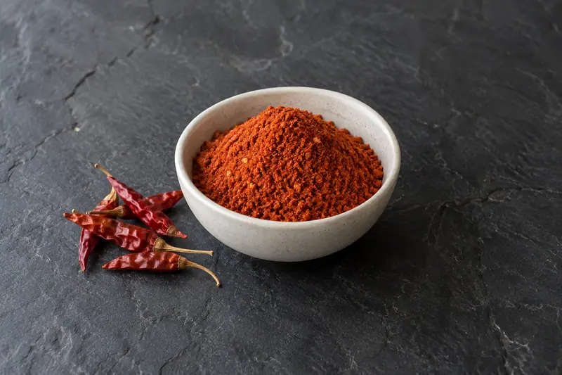 Orange-red chili powder heaped in a plain ceramic bowl beside dried bird’s eye chilies on dark slate.