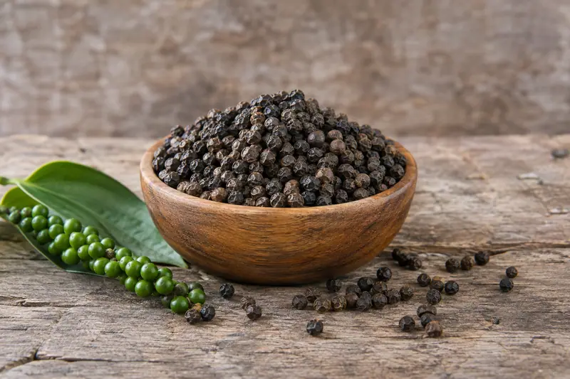 A wooden bowl heaped with black peppercorns, scattered peppercorns and fresh green pepper berries on a rough wooden table.