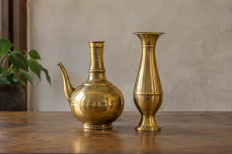 A polished brass kendi and tall flared vase stand on a wooden table against a neutral wall in soft daylight.