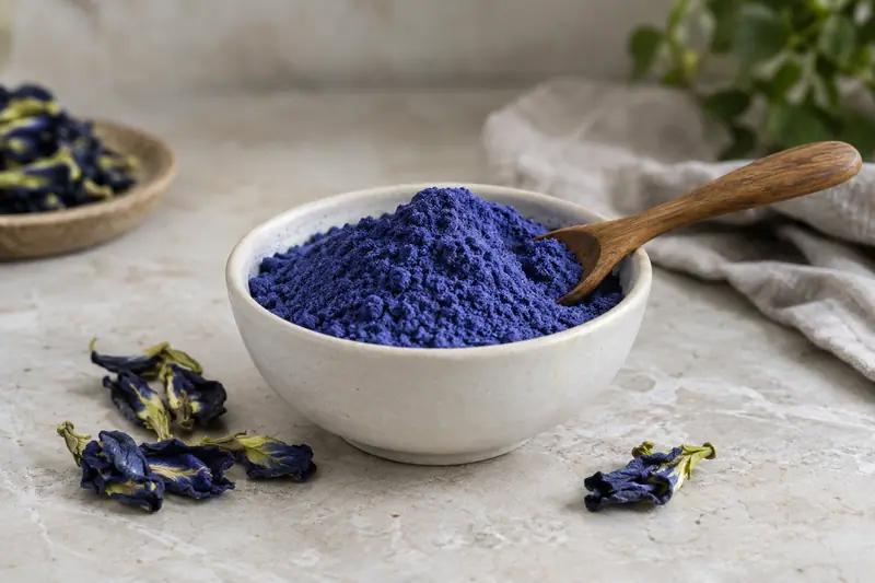 Deep blue butterfly pea powder in a ceramic bowl with a wooden spoon and dried flowers on pale stone.