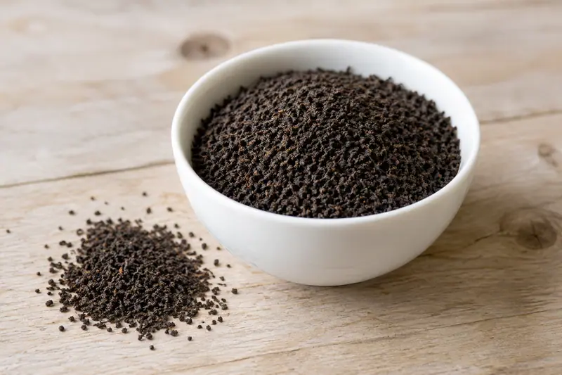 Fine dark tea granules fill a white porcelain bowl, with a small spread beside it on a pale wooden bench.