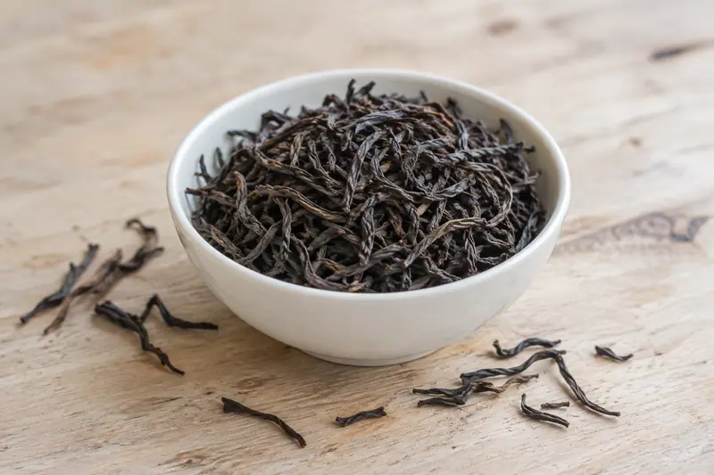 Long, twisted black tea leaves fill a white porcelain bowl on pale wood, with loose strands scattered beside it.