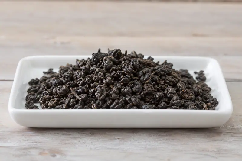 Tightly rolled dark tea leaves form a shallow heap on a white rectangular tray on a pale wooden bench.