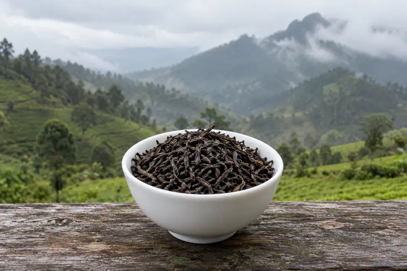 Dry black tea fills a white porcelain bowl on a weathered wooden ledge, with misty tea-covered mountains behind.