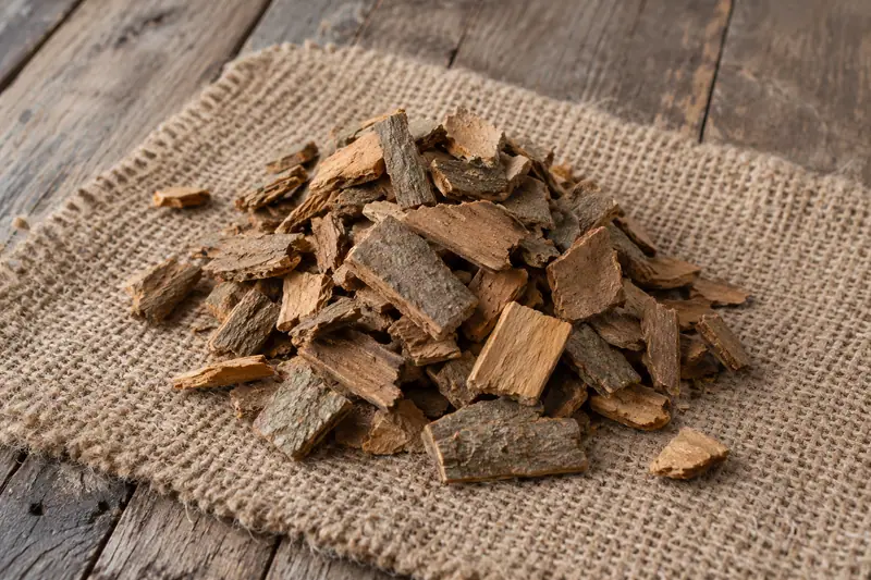 A heap of rough brown Ceylon cinnamon bark chips on jute cloth over a weathered wooden floor in natural daylight.