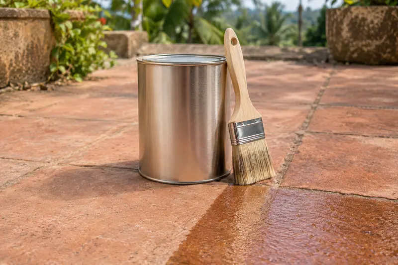 A plain metal tin and wide paintbrush stand on terracotta paving tiles beside a darker, glossy sealed section.