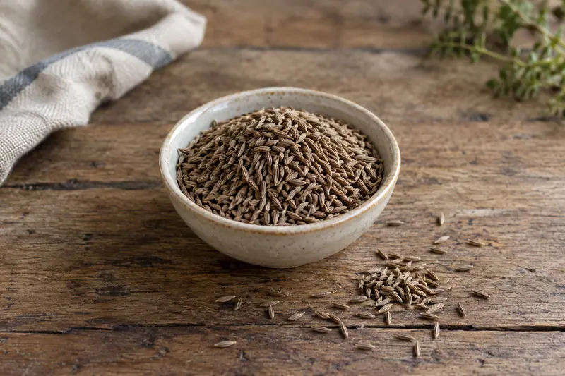 A plain ceramic bowl filled with cumin seeds sits on a rustic wooden table, with seeds scattered beside it.
