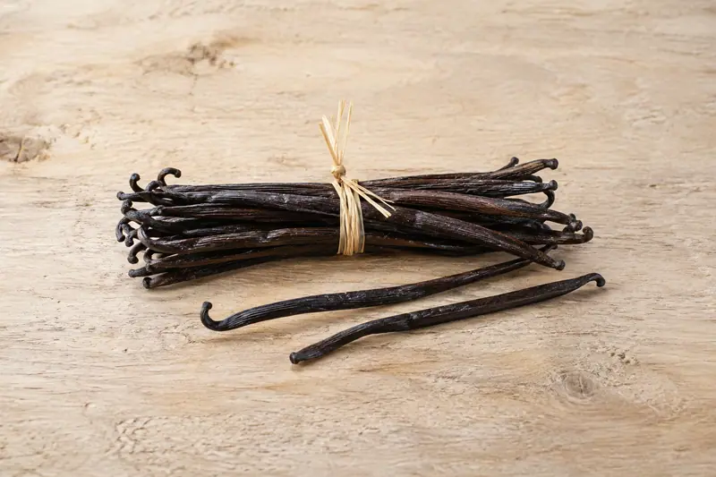 Dark cured vanilla pods tied with raffia rest on pale wood, with two loose pods beside the bundle.