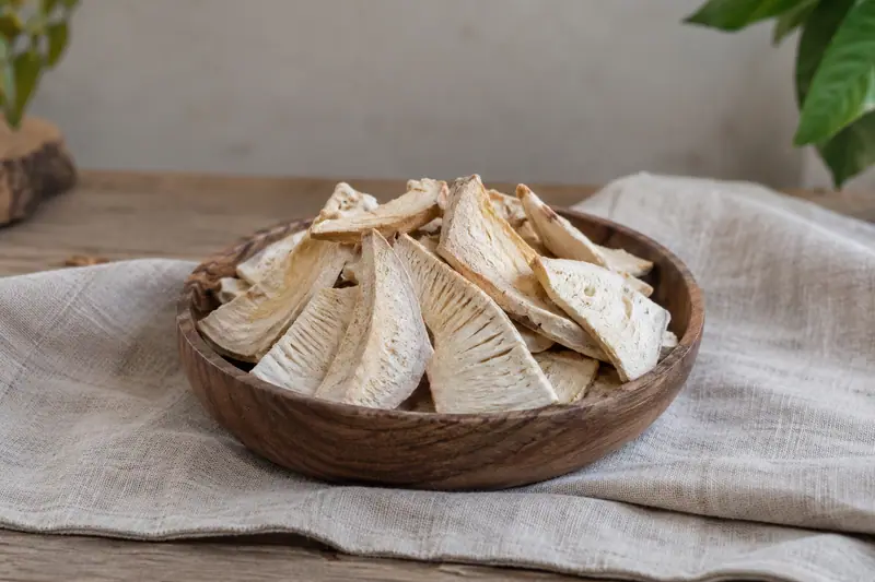 Pale dried breadfruit wedges piled in a shallow wooden bowl on light linen cloth in soft natural daylight.