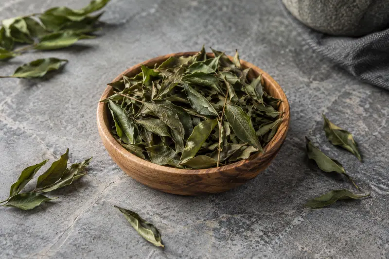 Dried curry leaves fill a shallow wooden bowl, with curled leaves and thin stems scattered on a grey stone surface.