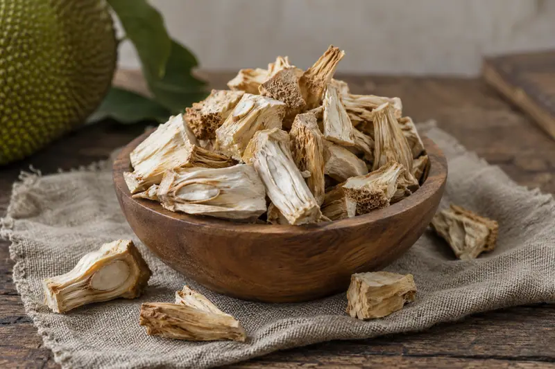Pale, fibrous dried young jackfruit pieces fill a wooden bowl, with a few scattered on rough linen cloth.