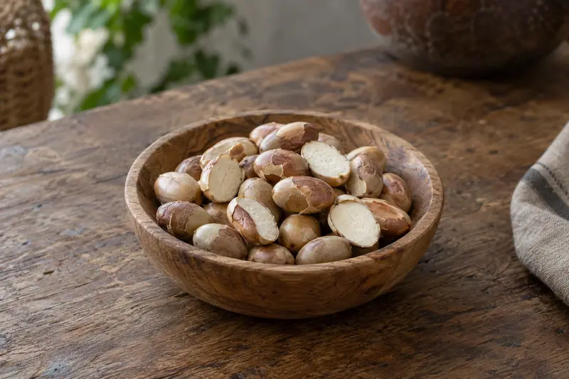 Whole and halved dehydrated jackfruit seeds with peeling brown skins in a shallow wooden bowl on a wooden table.
