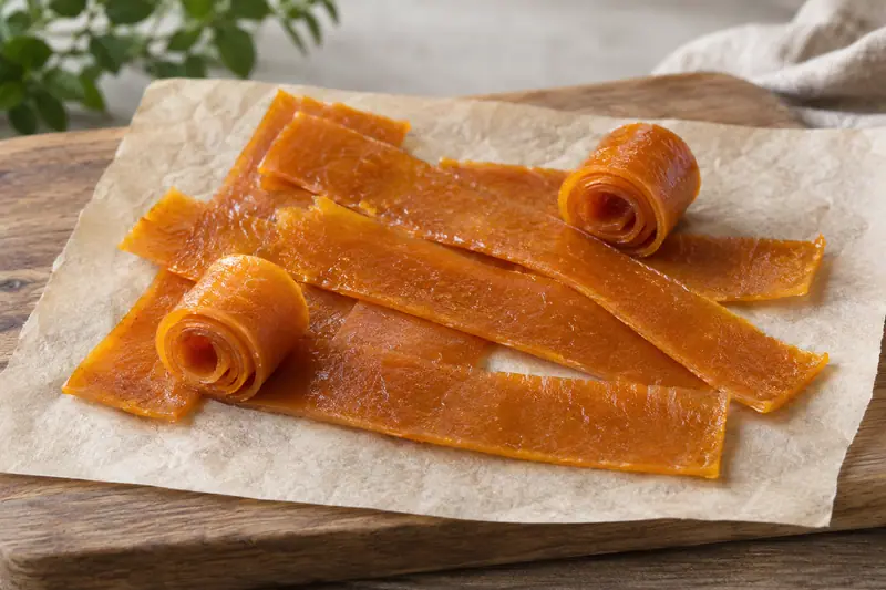 Amber-orange mango fruit leather strips and two rolls rest on plain baking paper on a wooden board in soft daylight.