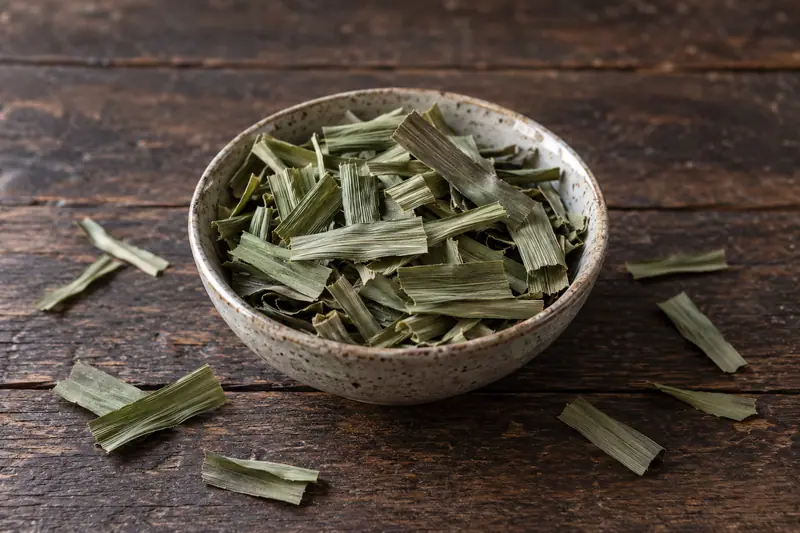 Dried olive-green pandan strips fill a shallow ceramic bowl, with scattered pieces on a dark wooden surface.