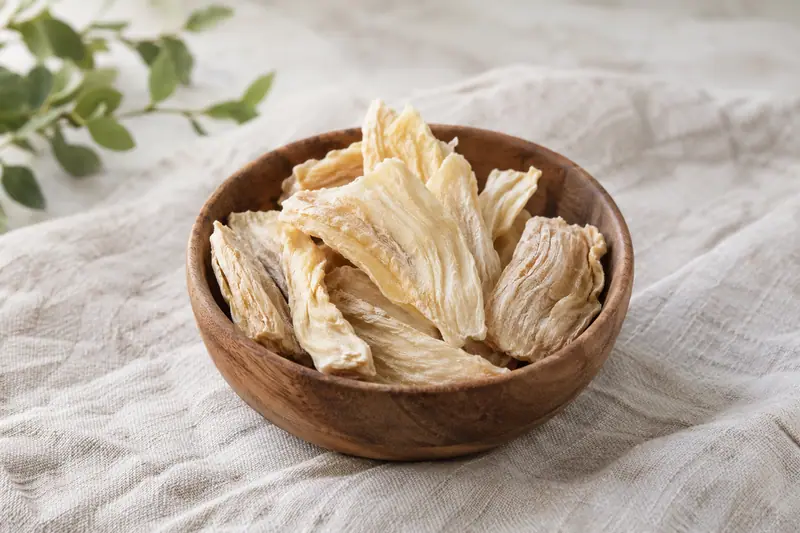 Pale, fibrous dried soursop pieces in a wooden bowl on cream linen in soft natural daylight.