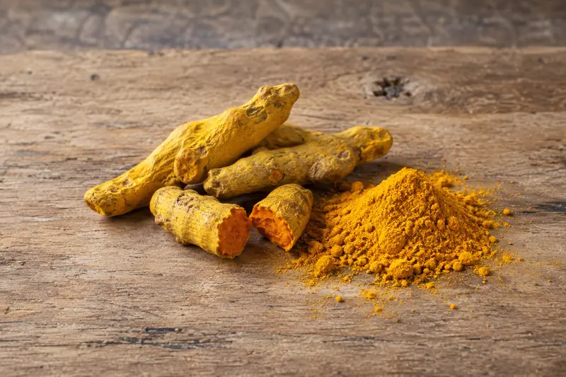 Dried turmeric fingers, one snapped to reveal its orange interior, beside golden turmeric powder on a wooden board.