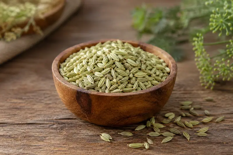 Pale green fennel seeds fill a wooden bowl, with a few scattered on a rustic wooden table in soft daylight.
