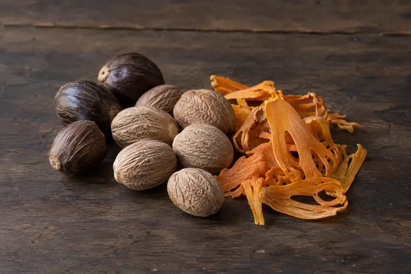 Light brown nutmeg kernels, dark unshelled nutmegs and orange dried mace rest on a dark wooden board.