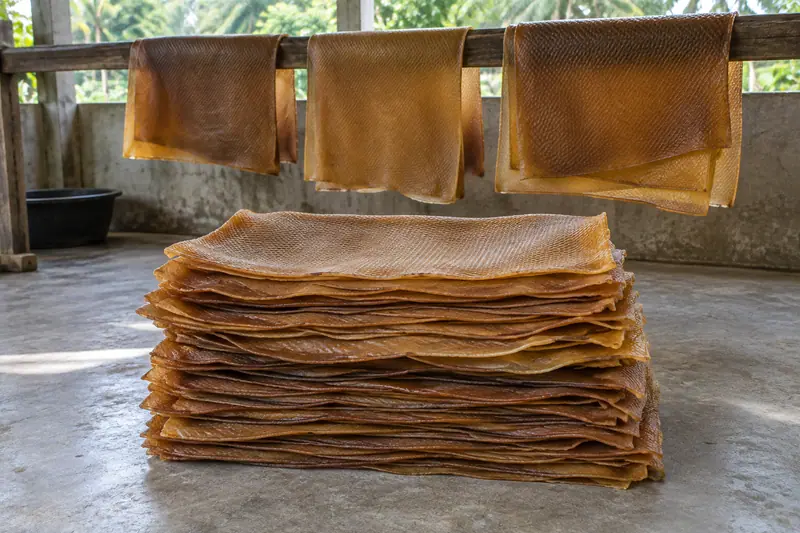 Amber-brown ribbed rubber sheets stacked on a concrete floor, with more sheets hanging over a wooden rail behind.