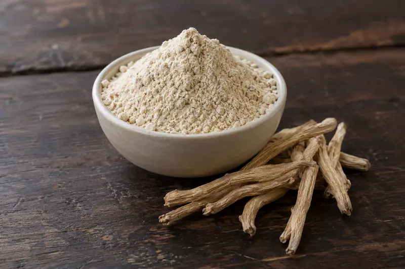 A ceramic bowl heaped with cream-beige shatavari powder sits beside dried tan roots on a dark wooden surface.