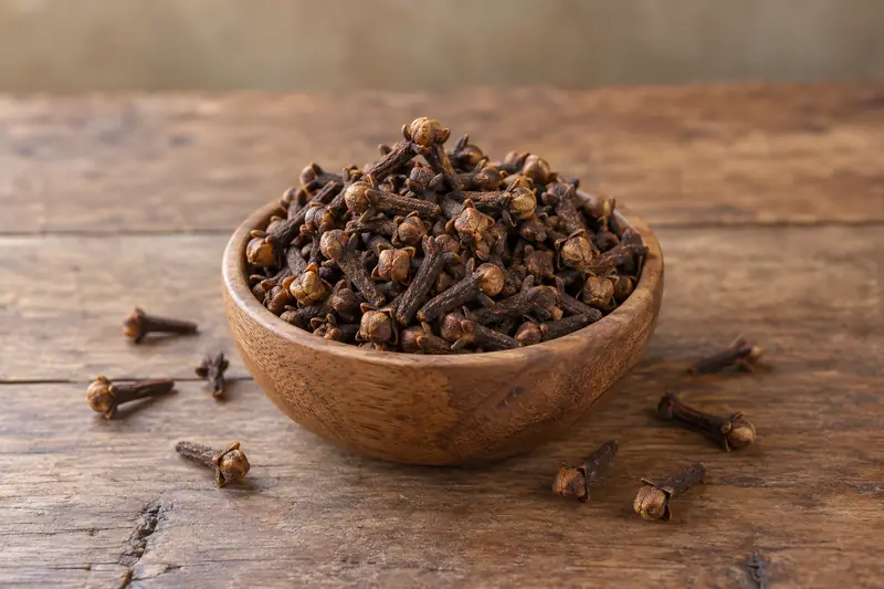 Whole dried cloves heaped in a small wooden bowl, with a few scattered on a rustic wooden table in soft daylight.